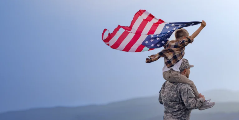 A soldier in camouflage holds a child on shoulders. The child waves an American flag against a blue sky.